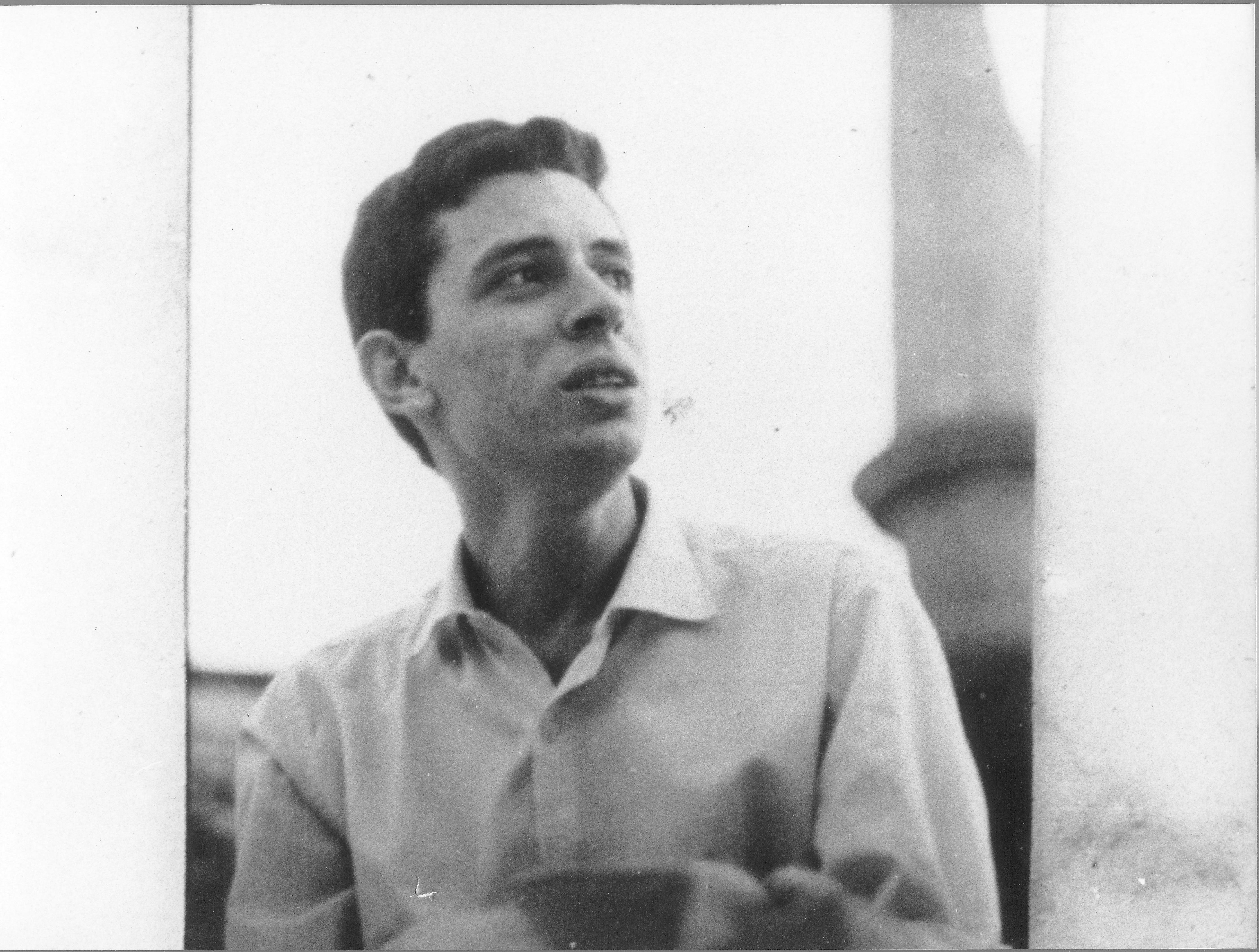 Homem jovem em foto preto e branca. Ele usa camisa branca e olha para o lado. Seus cabelos são pretos e curtos.
