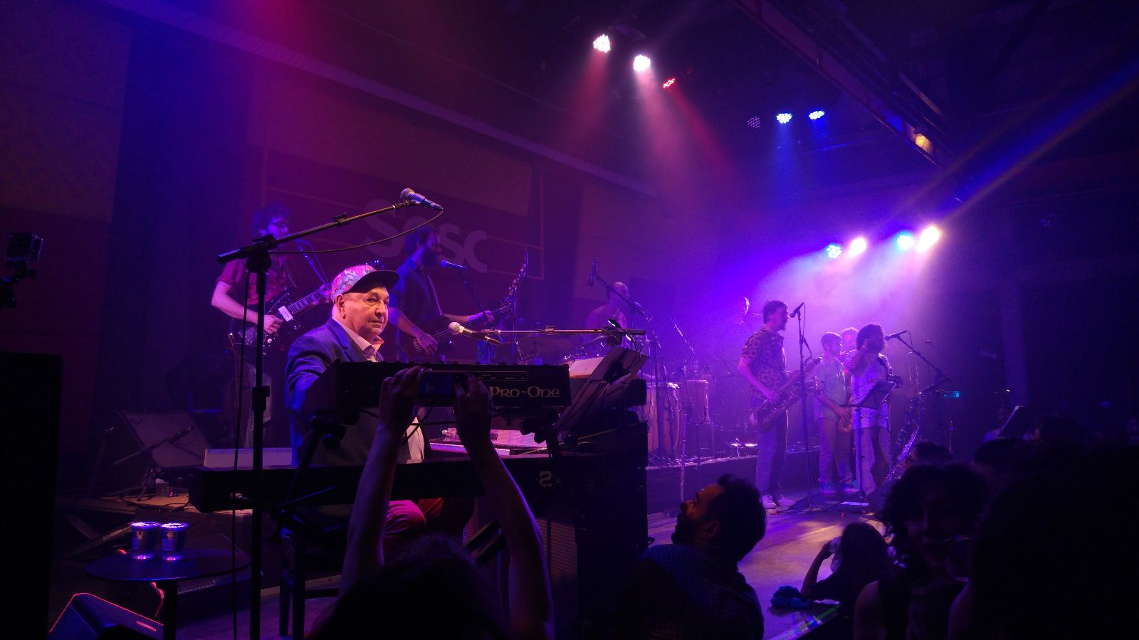 Fotografia de Jo&atilde;o Donato em apresenta&ccedil;&atilde;o. O artista est&aacute; do lado esquerdo da foto, em sentado em frente a um teclado. Uma luz azul ilumina o palco. Ele usa blazer preto, cal&ccedil;a vermelha e bon&eacute; colorido.