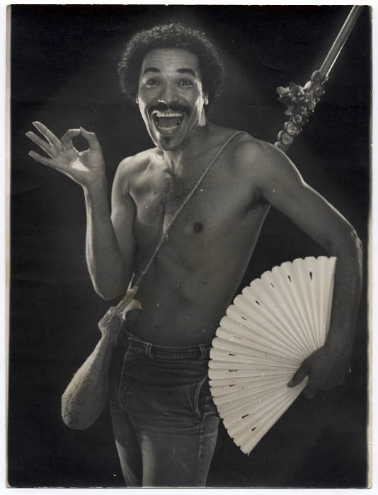 Na imagem, em preto e branco, está Marku RIbas. Ele posa sem camisa, sorrindo e fazendo sinal com uma das mãos. Na outra mão, ele segura um leque aberto. 