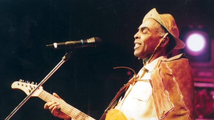 Gilberto Gil est&aacute; no palco, tocando viol&atilde;o e perto do microfone. Ele usa um chap&eacute;u de couro e uma capa.