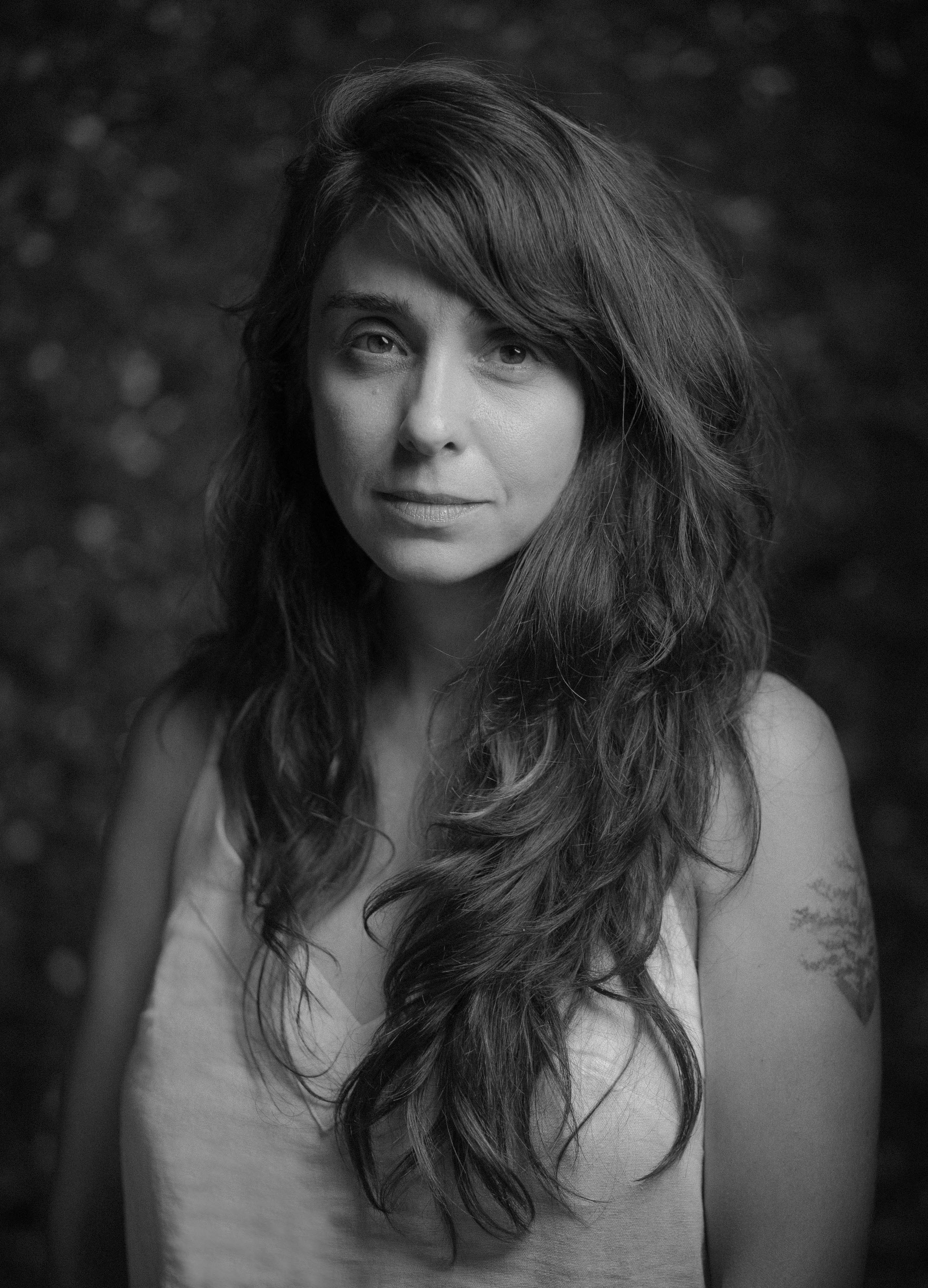 Mulher branca aparece em foto com os cabelos longos, repicados e lisos para a frente. Ela usa uma roupa branca de al&ccedil;a e tem uma tatuagem grande no bra&ccedil;o esquerdo.