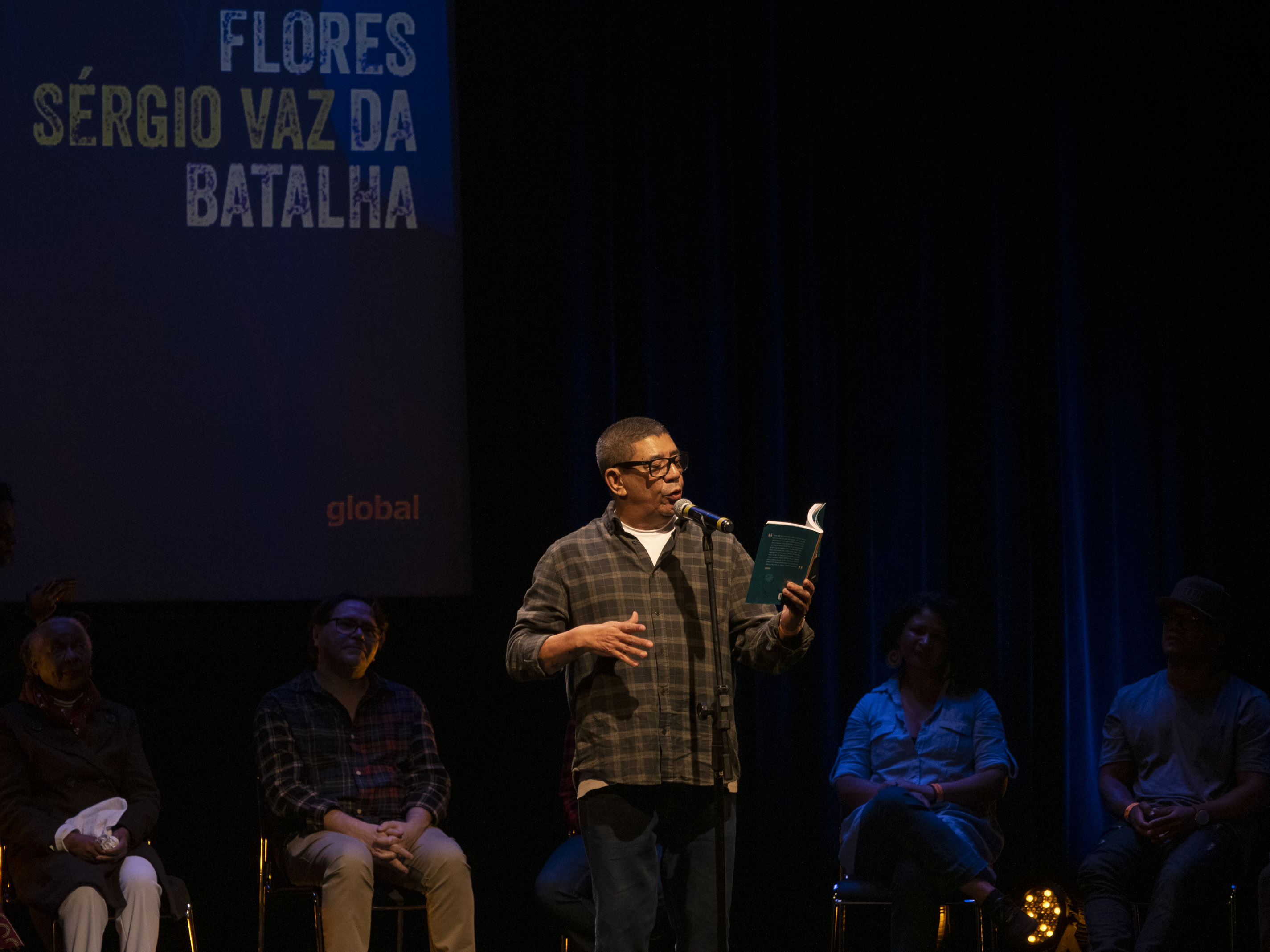 Sérgio Vaz está no palco, em pé, lendo. Ele usa óculos e veste uma calça jeans e uma camisa quadriculada.