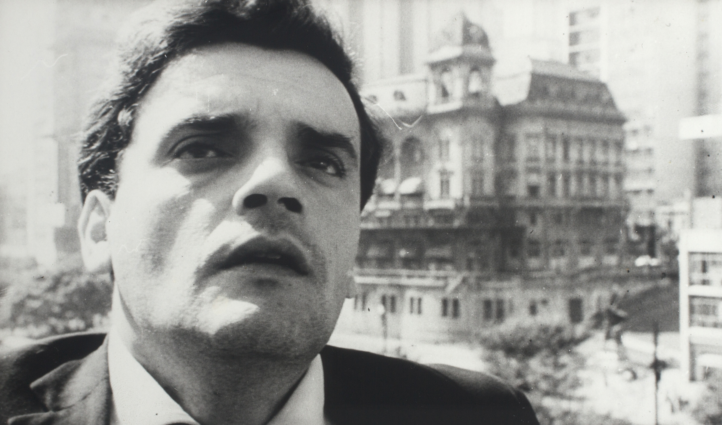 Imagem em preto e branco de cena do filme S&atilde;o Paulo Sociedade An&ocirc;nima. Na imagem, feita num plano fechado no rosto de um ator, h&aacute; um homem com um olhar de surpresa e ao fundo a cidade de S&atilde;o Paulo.
