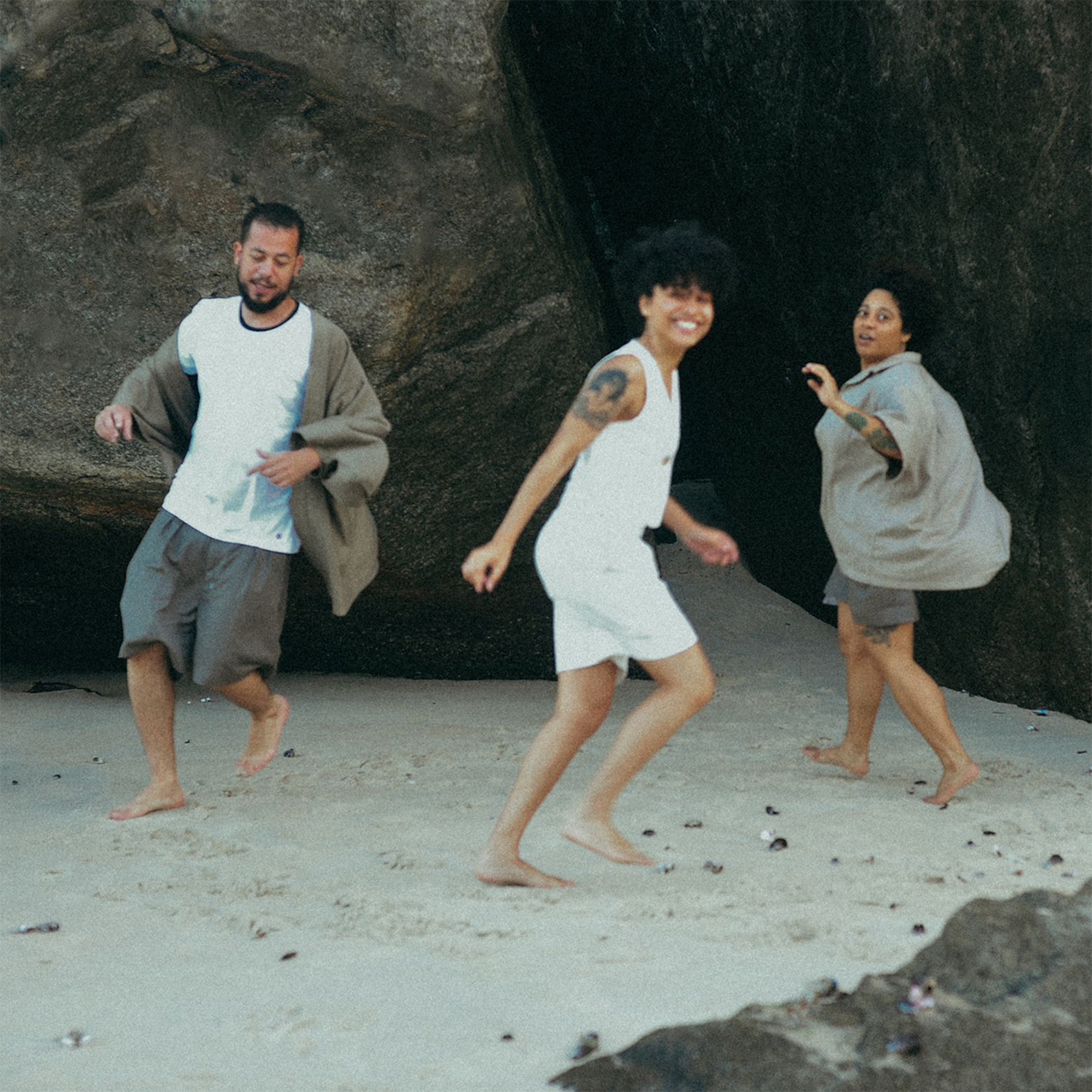 Fotografia colorida com os integrantes do grupo Tuyo caminhando em círculos na areia, descalços, próximos a uma grande pedra. Os integrantes são duas mulheres negras e um homem negro e todos vestem roupas casuais em tons neutros. 