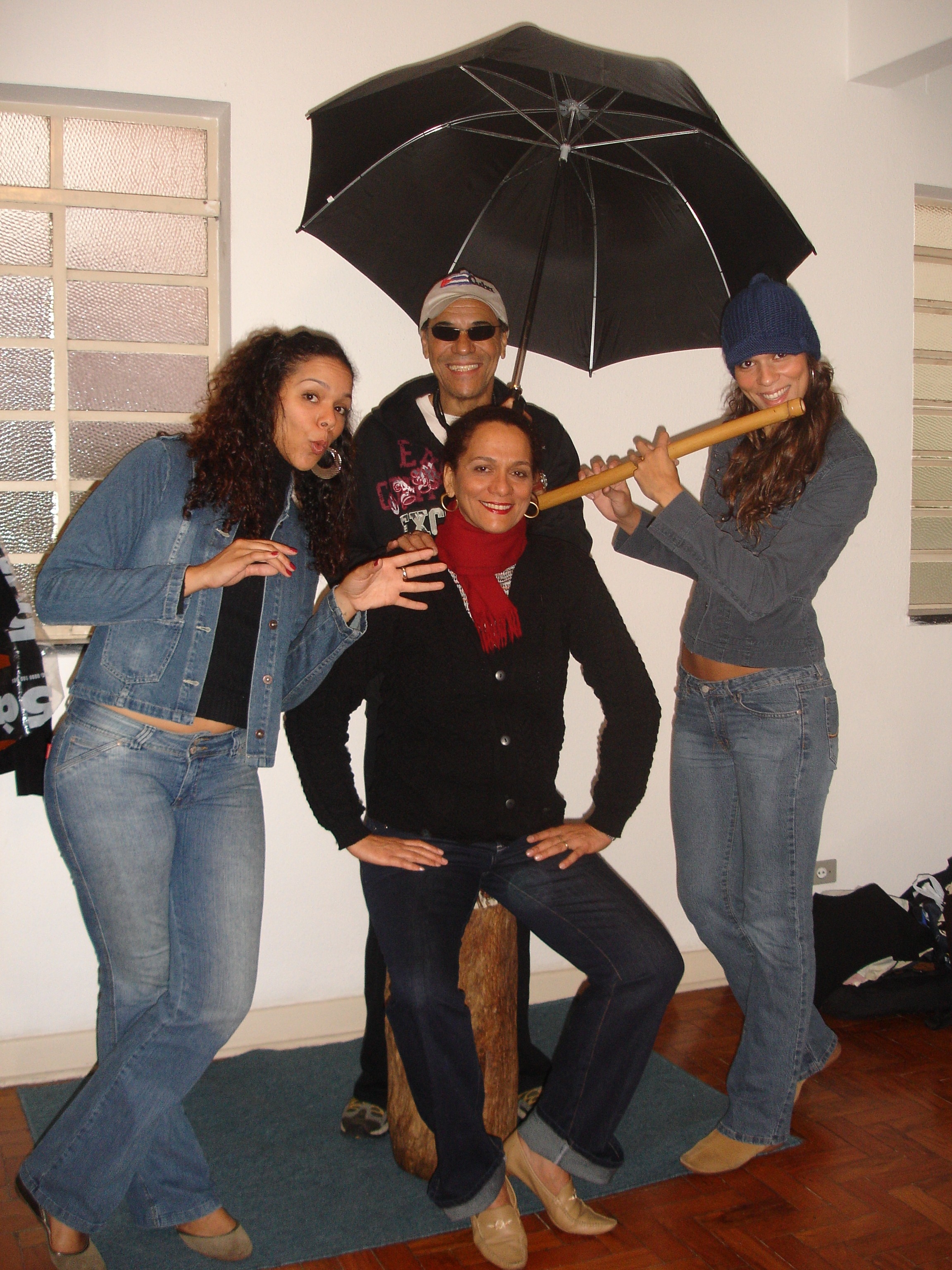 Há quatro pessoas na imagem, sendo três mulheres e um homem. A primeira está de pé e usa blusa preta, jaqueta e calça jeans. A do meio está sentada em um banco de madeira  usa blusa preta de magas compridas, lenço vermelho no pescoço e calca jeans. A terceira está tocando uma flauta e usa blusa azul e calça jeans. O homem, ao centro, segura um guarda chuva aberto, usa óculos escuros e blusa de frio preta. 