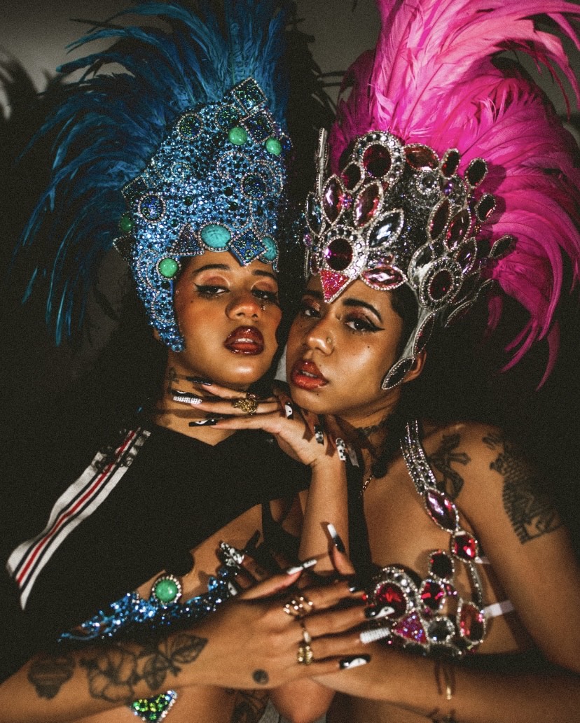 Fotografia colorida das irm&atilde;s gem&ecirc;as Tasha & Tracie, duo de hip hop. As duas irm&atilde;s s&atilde;o mulheres jovens, negras e est&atilde;o vestindo adere&ccedil;os coloridos na cabe&ccedil;a. Est&atilde;o pr&oacute;ximas uma a outra, lado a lado. 