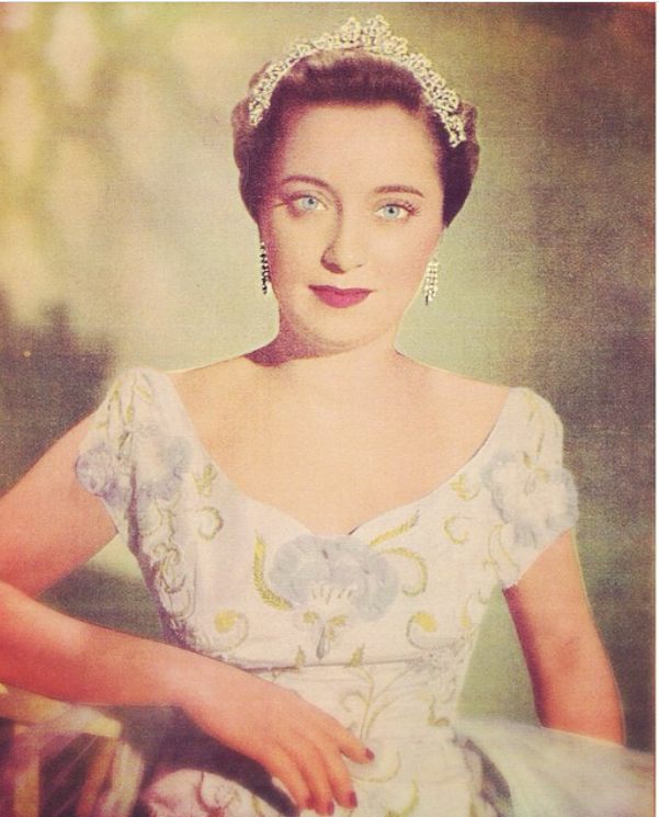 Mulher branca, de olhos azuis, usa vestido branco, com colo à mostra. Ela está com uma coroa e brincos grandes prateados.