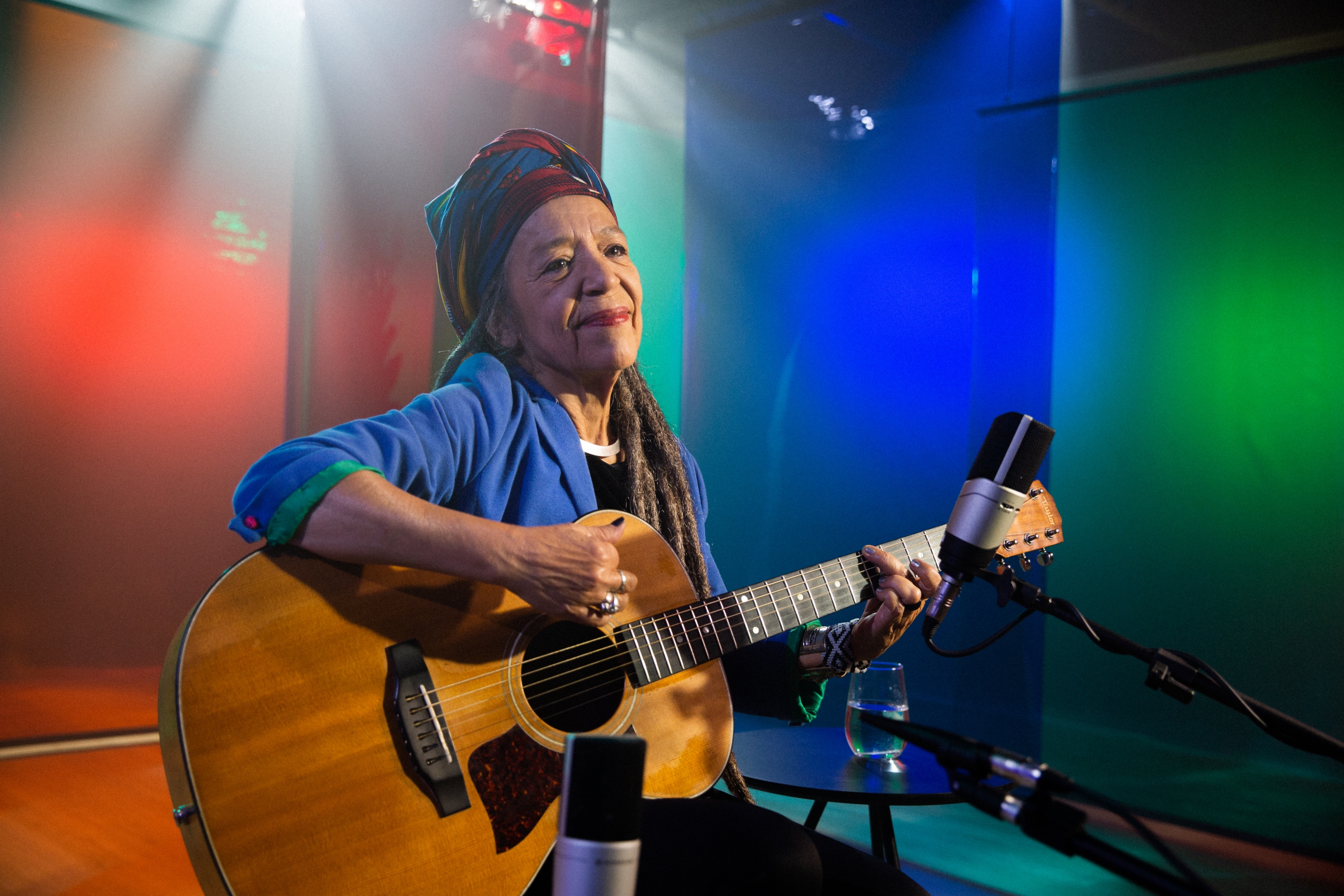 Na foto está uma mulher branca, de dreads nos cabelos, usando uma camisa azul e lenço colorido na cabeça. Ela está em um estúdio iluminado, com paredes nas cores verde, azul e vermelho. Ela está sentada em um banquinho e toca violão. Há um microfone próximo a ela.