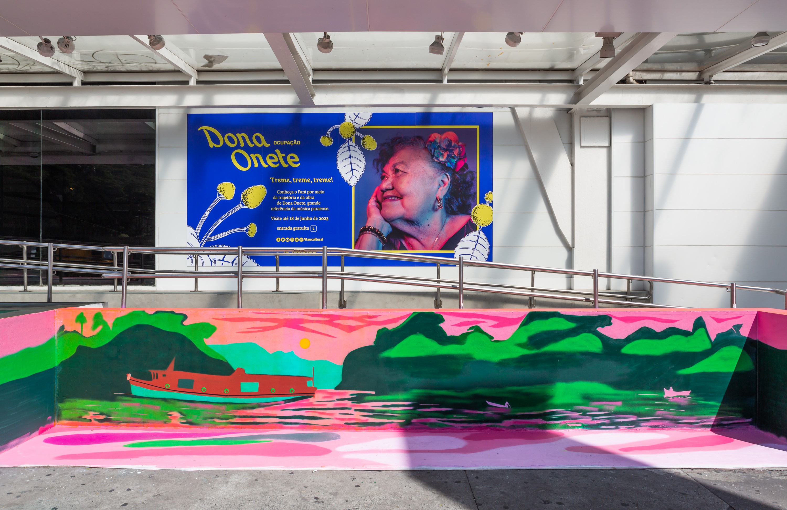 Fotografia colorida de um detalhe da pintura do artista visual Kamb&ocirc;. A obra foi feita no banco na fachada do Ita&uacute; Cultural. A imagem mostra um lado da pintura, focando em um barco navegando em um rio e a vegeta&ccedil;&atilde;o no entorno. As cores s&atilde;o verde, rosa, azul e vermelho.