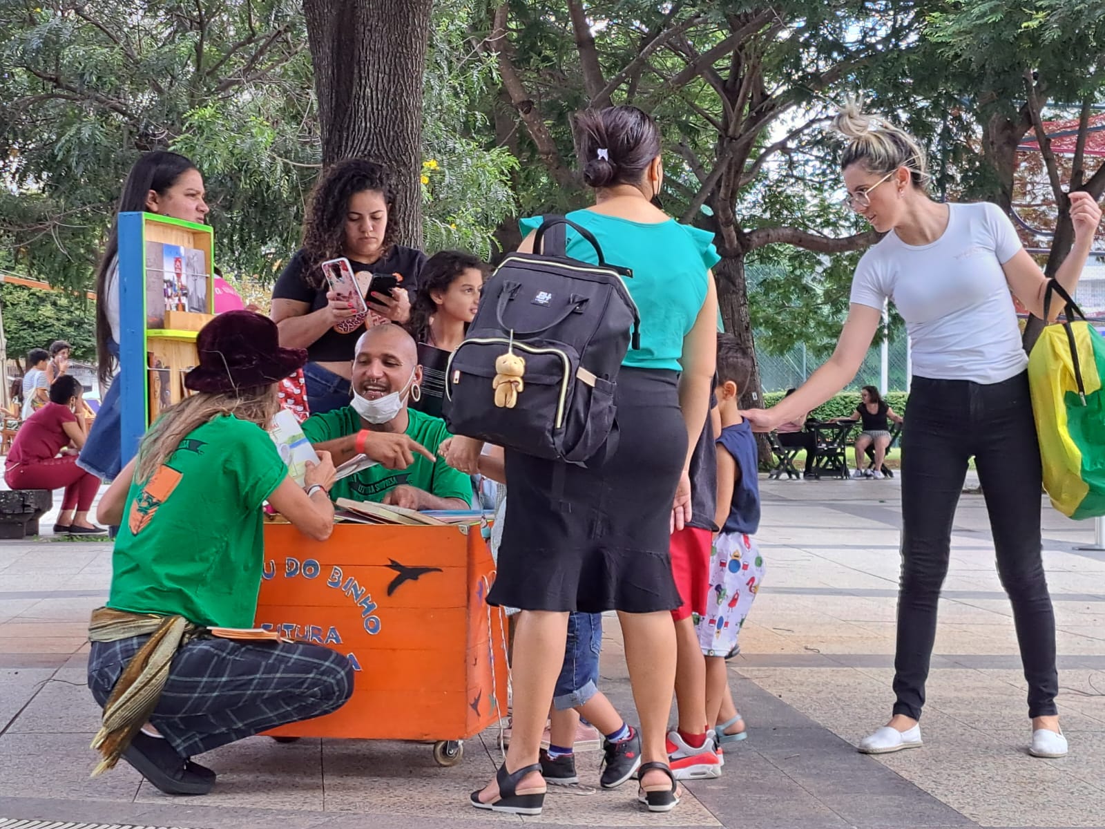 Na imagem há várias pessoas em uma calçada: homens, mulheres e crianças. Um homem e uma mulher, ambos com camisetas verdes, estão agachados e manuseando livros que se encontram dentro de uma caixa de madeira na cor laranja. Há pessoas ao redor e árvores ao fundo.