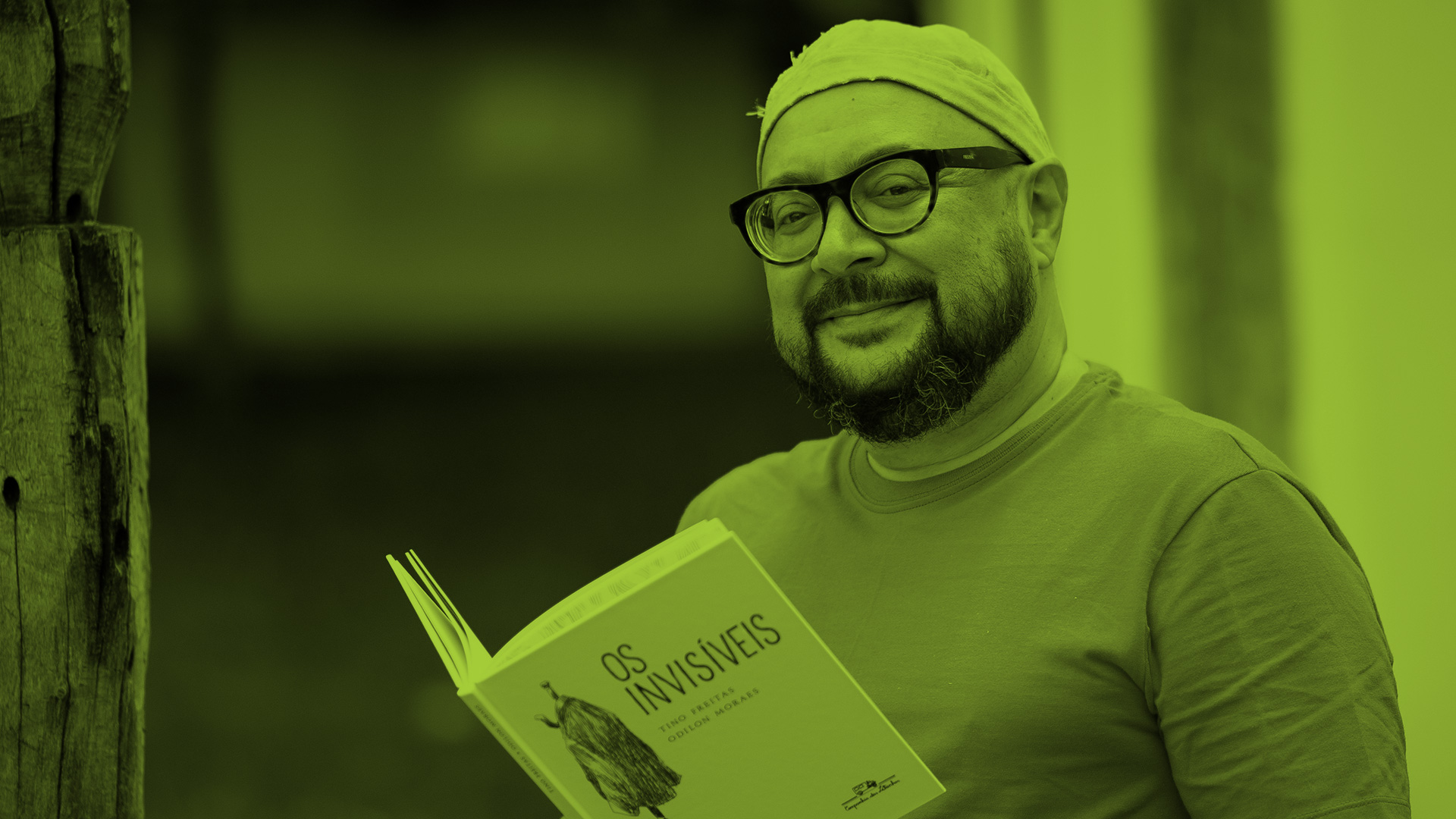Fotografia do autor Tino Freitas. Ele &eacute; um homem branco, com barba e &oacute;culos. Na foto est&aacute; de touca. Tino est&aacute; segurando um livro aberto. A imagem possui um filtro verde.