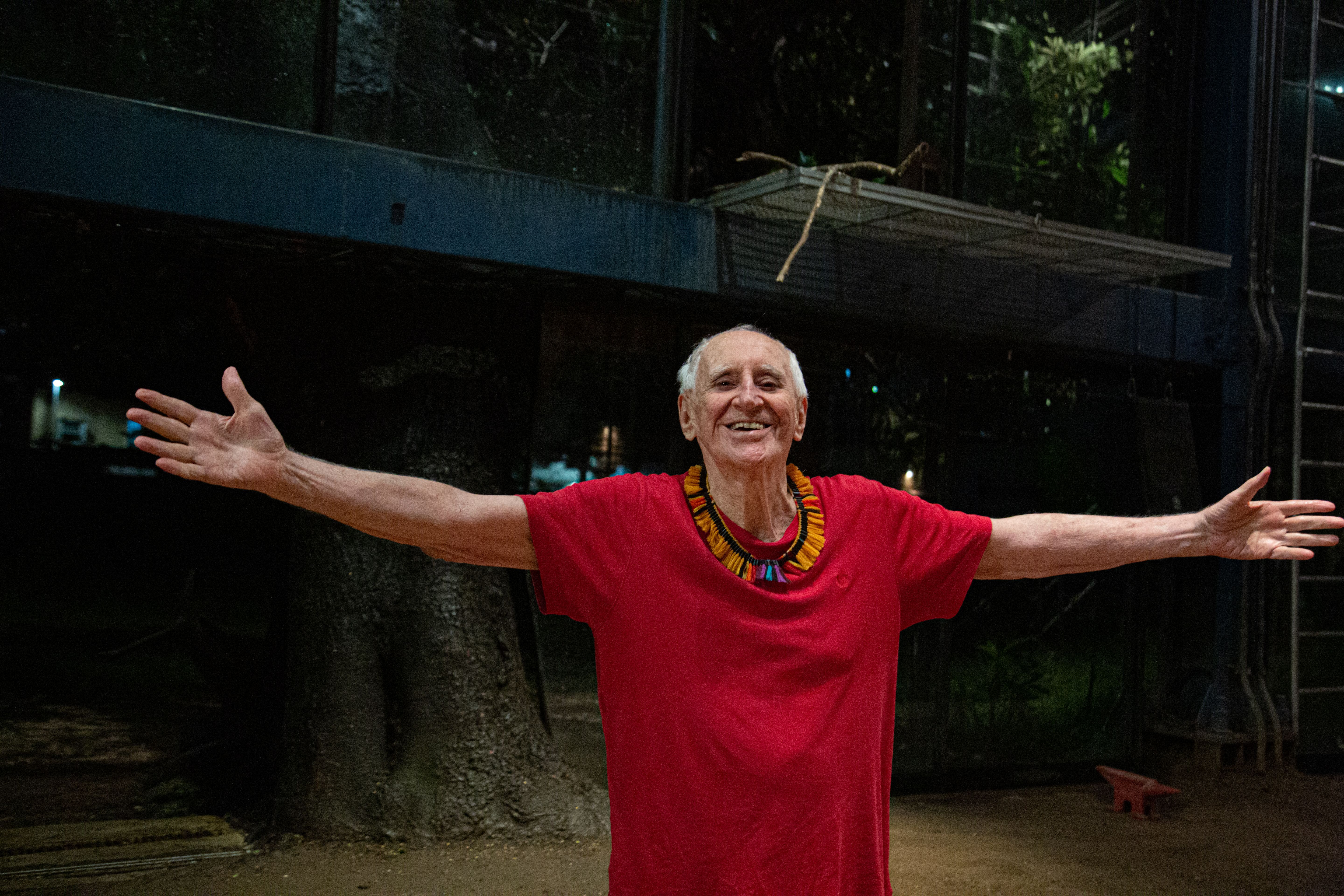 Homem idoso, de cabelos brancos, aparece de bra&ccedil;os abertos, sorrindo. Ele usa uma camiseta vermelha e um colar amarelo de mi&ccedil;angas.