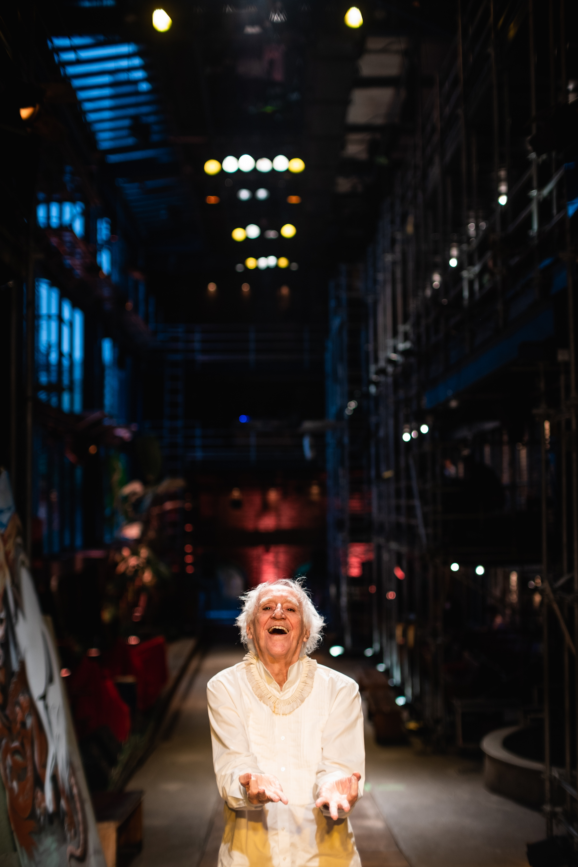 Homem idoso est&aacute; de p&eacute;, sorrindo, com as m&atilde;os abertas para a frente. Ele est&aacute; em um corredor relativamente estreito, com arquibancadas de ferro dos dois lados.