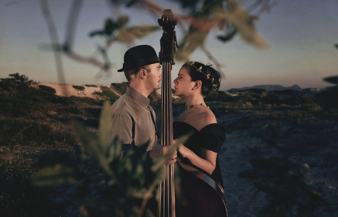 Fotografia colorida do duo A Corda em Si. Na imagem, Fernanda Rosa e Matheus Costa estão frente a frente, com um baixo acústico entre os dois. Ao fundo, uma paisagem com grama e areia. Fernanda usa vestido preto e um ramo de folha enrolado na cabeça e pescoço. Matheus veste camisa cinza e chapéu preto.