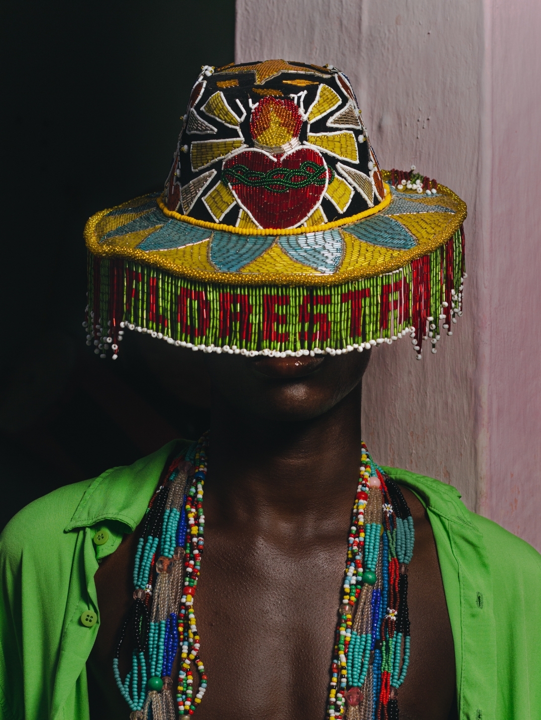 Homem negro está com uma camisa verde aberta, muitos colares coloridos e um chapéu todo bordado com miçangas coloridas, onde se lê a palavra Floresta na aba de franjas, cobrindo seu rosto.