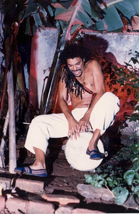 Na imagem está um homem de calça branca, faixa com franjas ao redor do corpo, com base no ombro, e sapatilhas azuis. Ele está sentado e tocando um instrumento. 