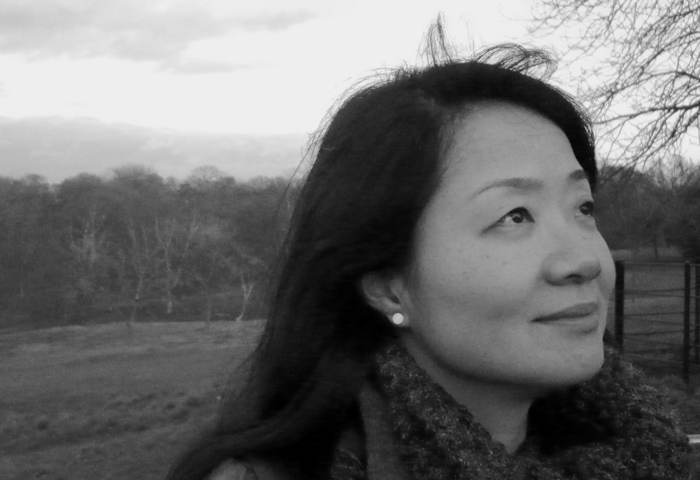 Mulher jovem, oriental, com cabelos pretos e lisos abaixo dos ombros, aparece em um campo ao ar livre. Ela est&aacute; olhando para cima, de cachecol, olhando levemente para cima.