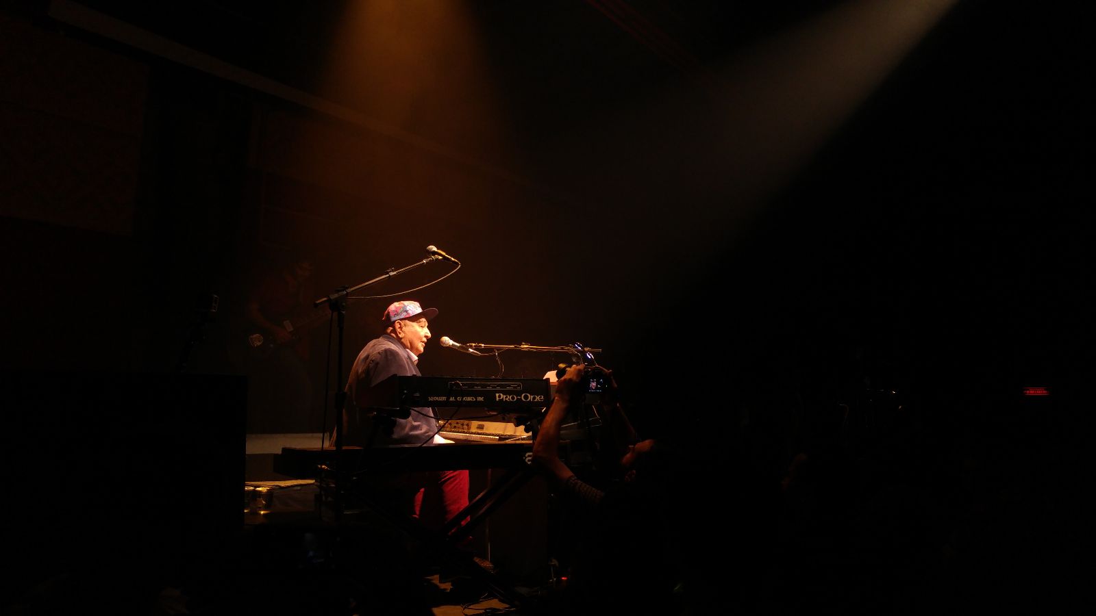 Fotografia de Jo&atilde;o Donato em apresenta&ccedil;&atilde;o. O artista est&aacute; do lado esquerdo da foto, em sentado em frente a um teclado. A luz do palco foca no artista enquanto todo o resto do palco est&aacute; escuro. Ele usa blazer preto, cal&ccedil;a vermelha e bon&eacute; colorido.