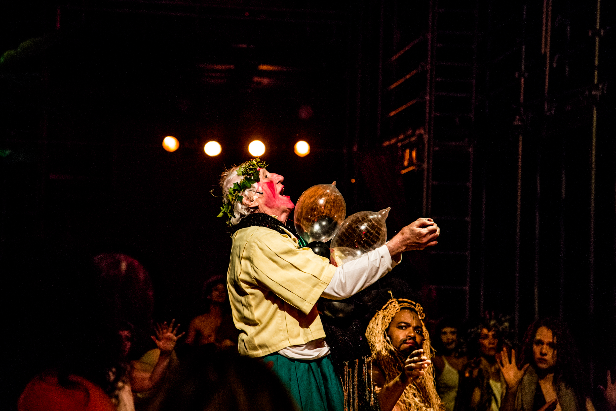 Homem idoso com enfeite de folhas na cabe&ccedil;a aparece em cena, com os bra&ccedil;os para cima, em um teatro. 