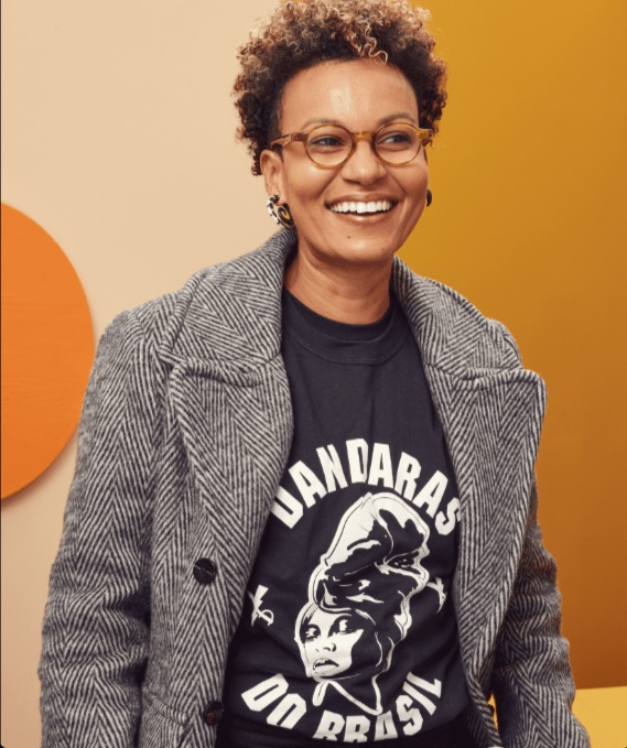 Mulher negra jovem aparece de p&eacute; e sorrindo. Ela usa &oacute;culos de grau, brincos pequenos estampados, blazer na cor cinza com listras pretas e brancas e uma camiseta preta por baixo, onde se l&ecirc; 