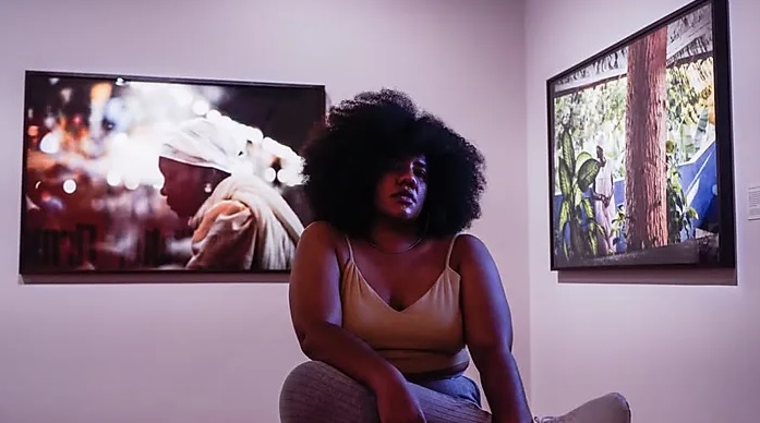 Mulher negra aparece sentada, com a perna cruzada e com os bra&ccedil;os em cima da perna. Ela tem os cabelos soltos na altura dos ombros, e usa uma blusa curta, de al&ccedil;a e na cor bege. Atr&aacute;s dela, pendurados em uma parede branca, h&aacute; duas fotografias. Uma delas mostra uma mulher negra com um len&ccedil;o branco na cabe&ccedil;a. 