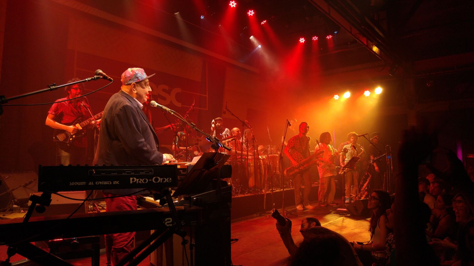 Fotografia de Jo&atilde;o Donato em apresenta&ccedil;&atilde;o. O artista est&aacute; do lado esquerdo da foto, em p&eacute; em frente a um teclado. Ele usa blazer preto, cal&ccedil;a vermelha e bon&eacute; colorido.