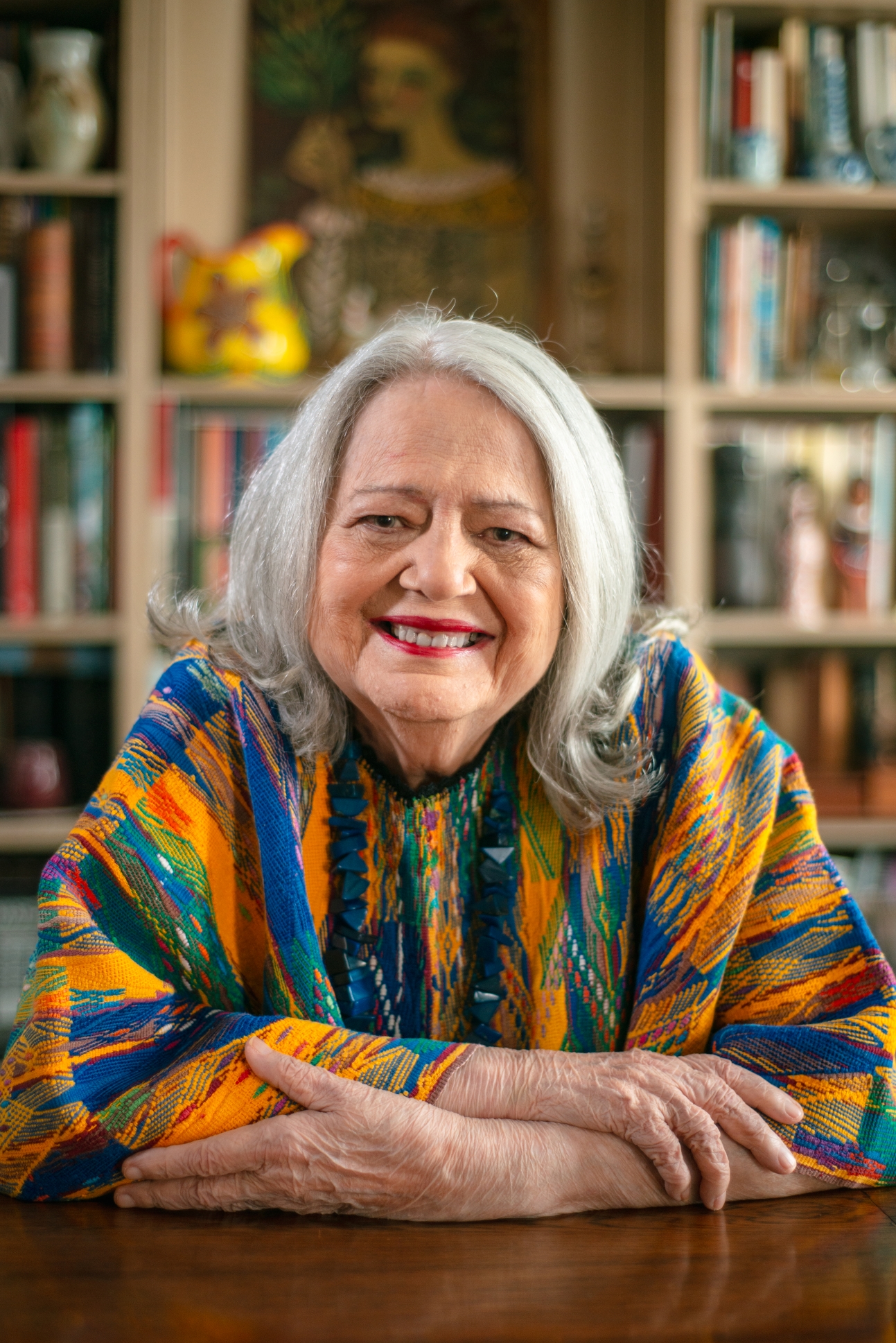 Retrato colorido da arte educadora Ana Mae Barbosa. Ela &eacute; uma mulher branca, com cerca de 80 anos de idade. Possui cabelos brancos, est&aacute; vestindo uma roupa colorida, com tons laranjas e azul. Olha para frente e est&aacute; sorrindo.