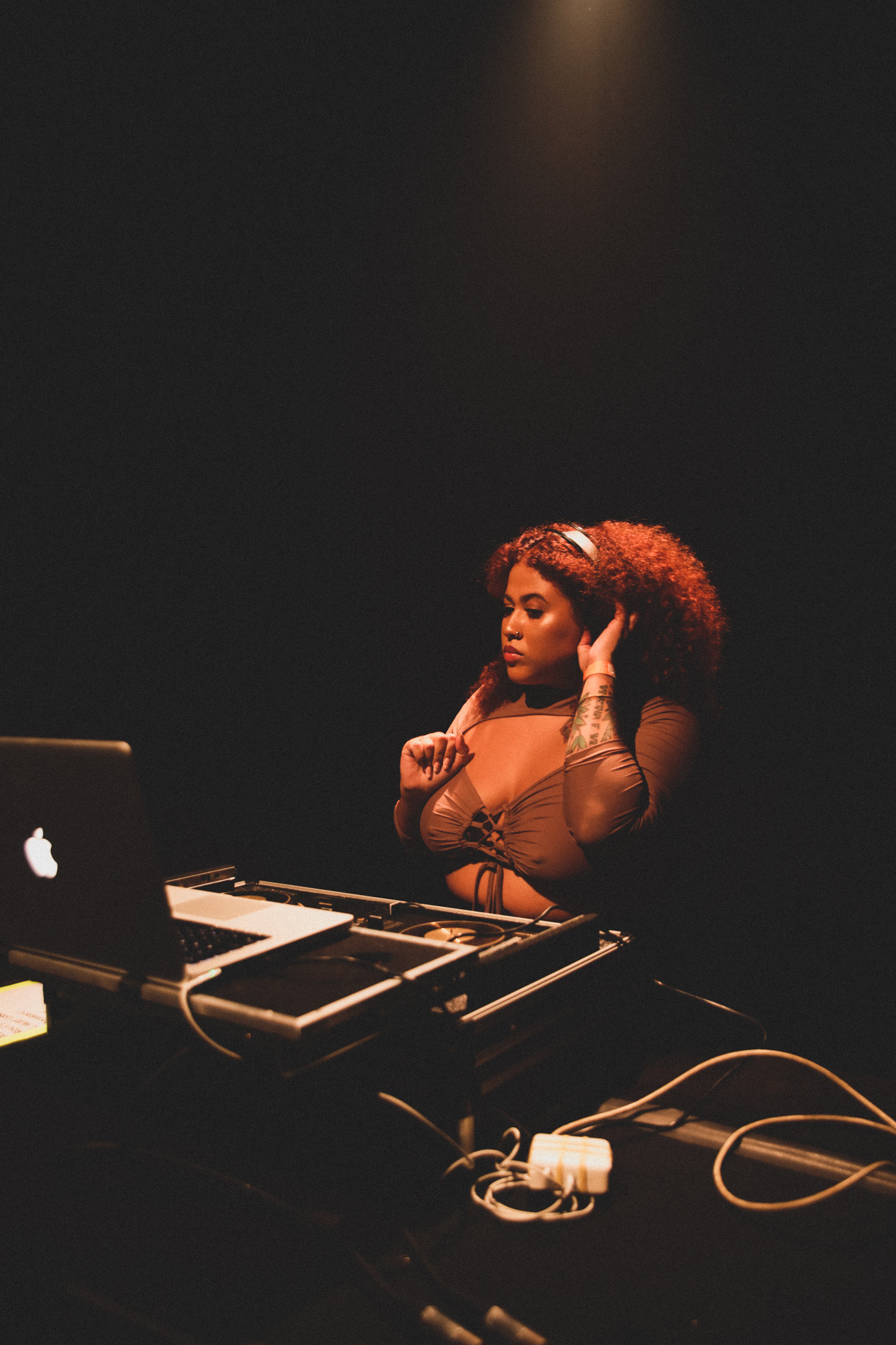 Fotografia colorida da DJ Livea. Ela &eacute; uma mulher jovem e negra, com cabelo cacheado e com uma tonalidade avermelhada. Est&aacute; em frente a uma mesa com equipamento sonoro, com fone de ouvido e um notebook em cima da mesa. 