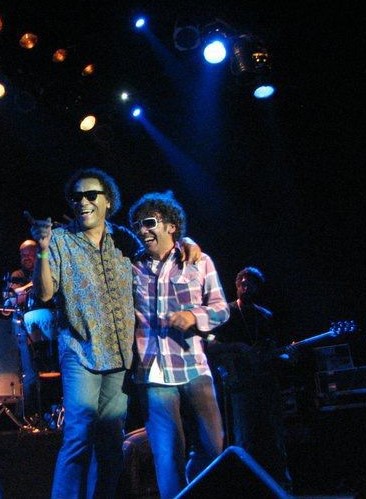 Dois homens posam juntos no palco. O primeiro, mais alto, usa óculos de sol, camisa estampada e está com um dos braços envolto no pescoço do outro. O segundo também usa óculos de sol, camisa xadrez e segura um microfone. 